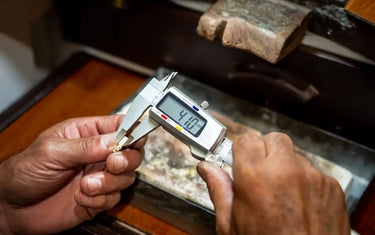 A jeweler measuring a small metal component at a bench with a digital caliper.
