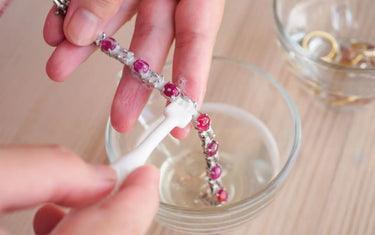 A person using a small soft brush to clean a silver ruby bracelet over a glass of cleaning solution.