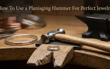 A planishing hammer resting on a jeweler bench pin next to a hammered silver ring, bracelet, and anvil.