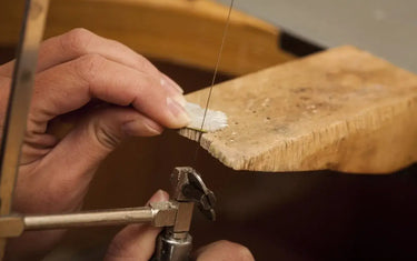 Close-up of a jeweler using a piercing saw to cut a precise design into metal on a wooden bench pin.