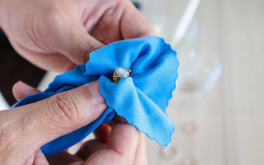 Close-up of hands cleaning a gold gemstone ring with a soft blue polishing cloth.