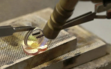 Close-up of soldering a sterling silver ring band using a blowtorch and tweezers on a soldering block.