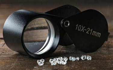 A professional black 10X-21mm folding jewelry loupe lying next to uncut rough diamond crystals on a wooden surface.