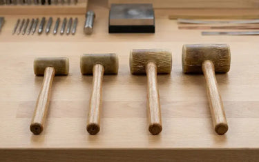 Four different sizes of rawhide mallets lined up on a wooden jewelry-making workbench.