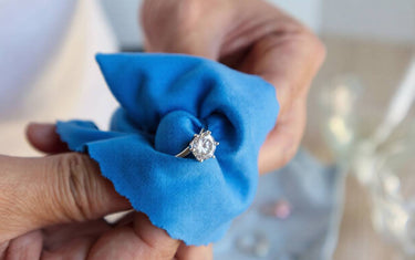 Close-up of hands polishing a silver diamond engagement ring with a blue microfiber jewelry cleaning cloth.