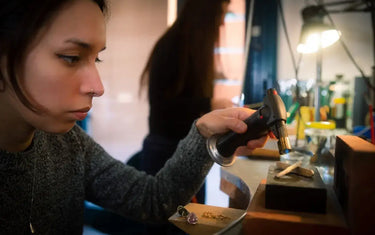 A female jeweler focuses intently on soldering a metal piece using a handheld butane torch at her studio workbench.