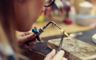 A jeweler wearing glasses uses a micro torch and tweezers to solder a small, intricate gold jewelry component on a soldering block.