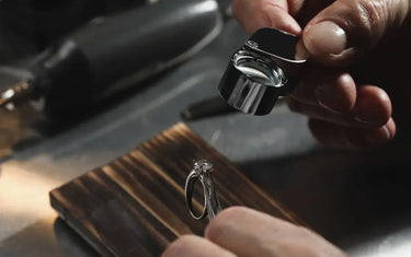 Close-up of a jeweler holding a folding loupe magnifier to inspect a diamond ring on a wooden bench pin.