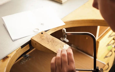 A jeweler using a hand saw frame to cut metal on a wooden bench pin at a jewelry making workstation.