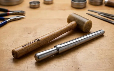 Rawhide mallet and steel ring sizer mandrel lying on a wooden jewelry-making workbench.