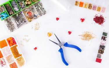 Flat lay of blue round-nose pliers surrounded by organized organizer boxes containing colorful jewelry beads and pins.