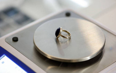 Close-up of a dark blue faceted gemstone set in a silver ring, resting on the metal weighing pan of a digital jeweler's scale.