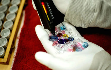 A person wearing white gloves uses a handheld electronic tester on a pile of colorful gemstones.