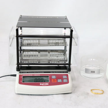 The MZ-K3000 tester balance positioned next to a glass beaker and a gold bracelet on a workspace.
