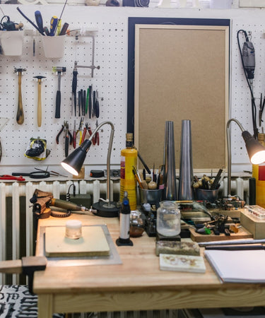 A jeweler's workbench with various tools hanging on a pegboard wall, desk lamps, and metal mandrels on the work surface, used for jewelry making and repair.