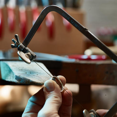 Close-up of a jeweler holding a metal ring band on a wooden bench pin while cutting it with a NIQUA piercing saw.