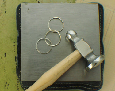 A steel bench block resting on a leather sandbag with silver jewelry rings and a chasing hammer.